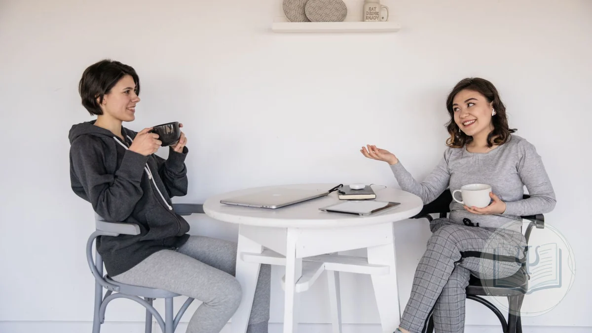 Dues persones conversant de manera relaxada en una taula, una escoltant i l'altra expressant-se amb un gest: comunicació assertiva i habilitats socials