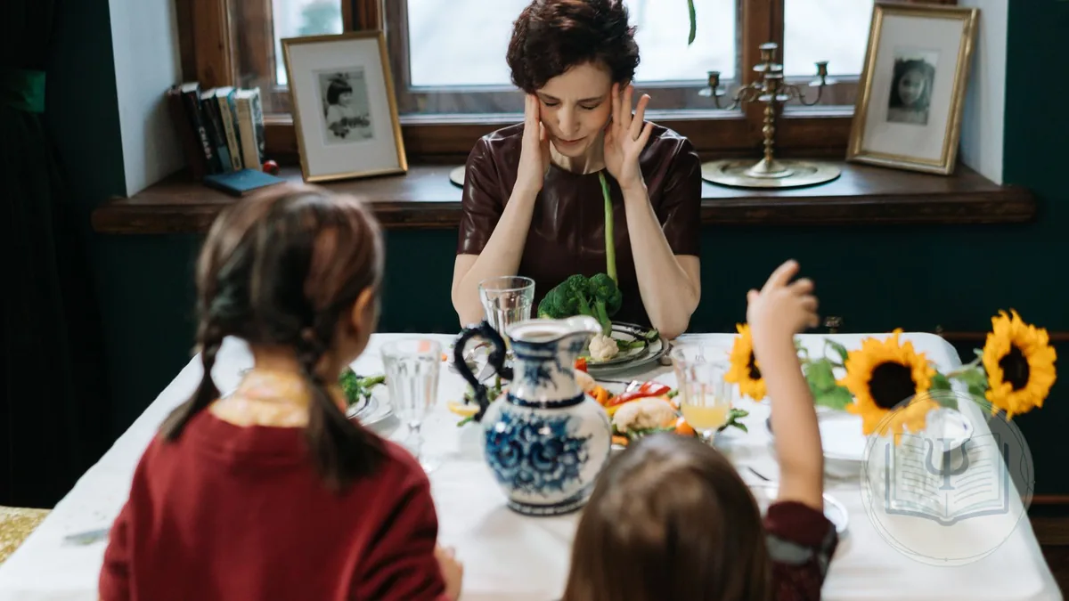 A woman sitting at the dining table with her hands on her temples and her eyes closed while her two daughters eat, a typical misophonia scene
