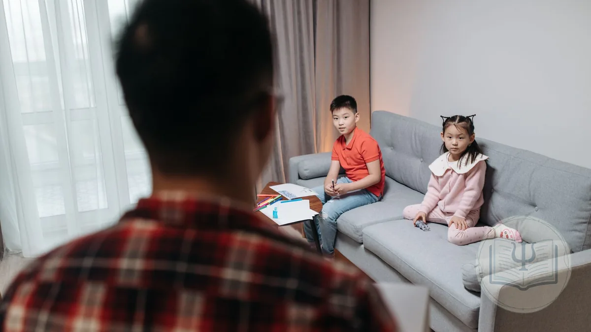 Systemic therapist seen from behind facing two siblings sitting on the consulting room sofa, with drawings and markers on the table